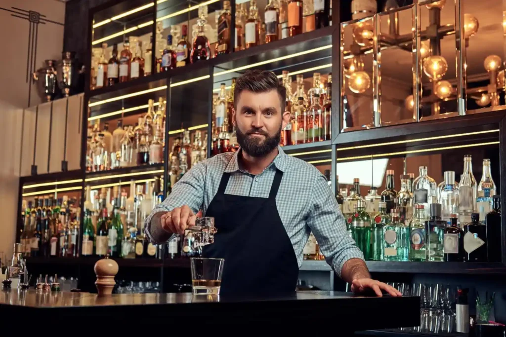 Professional bartender preparing a premium cocktail behind a fully stocked bar with liquor bottles and modern lighting